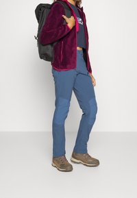 Chaqueta de forro polar en burdeos con detalles en rosa, combinada con pantalones azules con parches en las rodillas, y botas de senderismo marrones con cordones y suelas de goma.