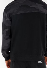 Czarny sweatshirt z teksturowanym tyłem w ciemne wzory kamuflażowe, zakończony ściągaczami oraz metką na dolnym brzegu. Materiał wydaje się miękki i trwały.