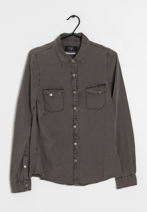 Chemise gris foncé à manches longues avec boutons sur le devant, deux poches poitrine, boutons visibles et col, suspendue sur un cintre noir sur fond blanc.