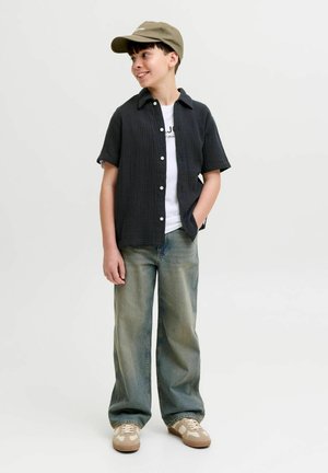 Dječak nosi kaki kapu, crnu majicu kratkih rukava preko bijele majice, široke izblijedjele traperice i bež tenisice, stoji sa rukom u džepu.