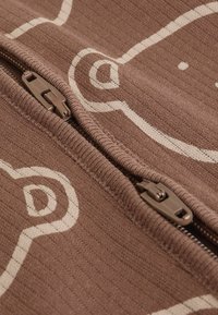 Primer plano de tela marrón con patrón abstracto beige con dos tiradores de cremallera de plástico marrón en una cremallera parcialmente abierta.