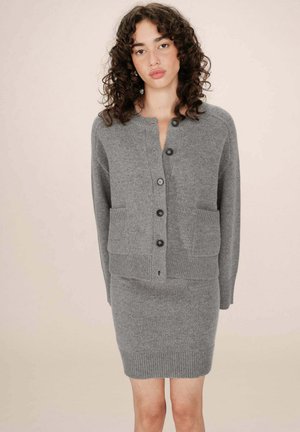 Femme aux cheveux bouclés portant un cardigan gris boutonné et une jupe en tricot assortie sur un fond beige uni.