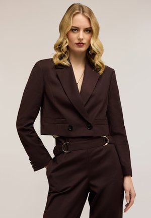 Femme blonde portant un blazer cropé double boutonnage marron foncé et un pantalon taille haute assorti avec une ceinture à boucle dorée.