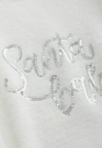 Tissu blanc avec les mots 'Santa' et 'Baby' en texte scintillant argenté, sur un fond doux et texturé et un design de lettres lisses et courbes.