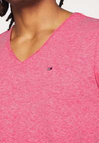 Rosa melerad bomullströja med V-ringning, prydd med en liten broderad logotyp på bröstet. Mjuk textur och avslappnad passform.