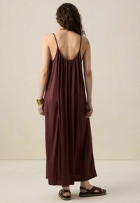 Robe maxi en tissu bordeaux foncé, avec des bRetelles fines réglables, un dos froncé et une coupe ample. Associée à des sandales marron.
