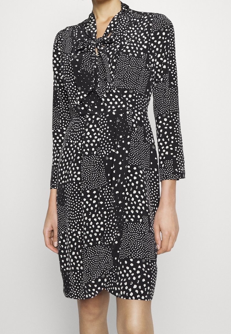 Svart klänning med vita pricken och mönsterdesign, försedd med knytkrage, långärmade och en figursydd midja för en strukturerad silhuett.