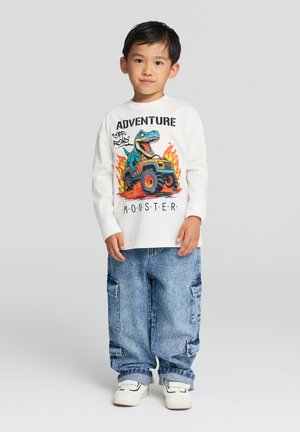 Camicia bianca a maniche lunghe con una grafica colorata di un dinosauro e di un monster truck con fiamme. Abbinata a jeans blu a vestibilità ampia e sneakers bianche.
