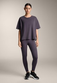 Žena stojí v uvoľnenej póze, oblečená v tmavo fialovom oversized tričku, zodpovedajúcich legínach a čiernych teniskách, na jednofarebnom béžovom pozadí.