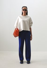 Sweatshirt oversized branco, calças largas azuis-marinho, mala laranja e ténis pretos com detalhes azuis. Óculos de sol e brincos de argola.