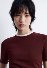 Burgundy ribbed t-shirt with a cream-colored round neckline, featuring short sleeves and a fitted design. Smooth texture, minimal detailing.