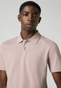 Ljust rosa pikétröja med krage, korta ärmar och en subtil logotyp på bröstet. Texturen ser jämn ut och tyget är andningsbart.