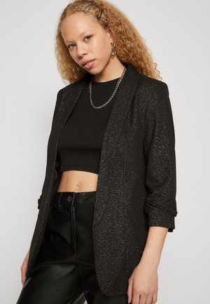 Junge Frau mit lockigem Haar trägt einen glitzernden schwarzen Blazer, schwarzen Crop-Top, Lederhosen, Kettenhalskette und Ohrringe vor einfarbigem Hintergrund.