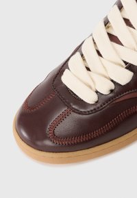 Primer plano de una zapatilla de cuero marrón con cordones color crema y costuras en zigzag visibles sobre una suela de goma.