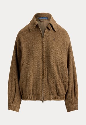 Personne portant des lunettes de soleil noires, une veste marron texturée, une chemise rouge, un pantalon blanc à jambes larges et des chaussures marron pointues, debout les bras croisés.