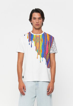 Young man wearing white T-shirt with colorful abstract dripping pattern and text on right shoulder, paired with light blue jeans.