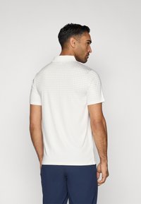 Balta polo krekls ar teksturētu punktiņu rakstu, īsām piedurknēm un apkakli; kombinēts ar tumši ziliem biksēm; redzams no aizmugures.