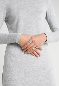 Mehrere Ringe an den Fingern, die verschiedene Designs zeigen: einer mit einem großen Stein, andere mit kunstvollen Mustern und kleinen Akzenten. Graues Langarmshirt.