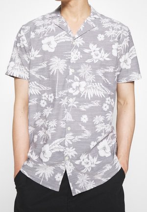 Homme portant une chemise grise à manches courtes boutonnée avec un motif tropical blanc de fleurs et de palmiers, mains dans les poches d’un pantalon noir.