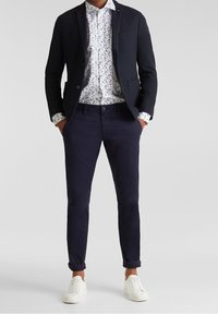 Blazer bleu marine, chemise blanche à motifs, et pantalon bleu marine, associés à des baskets blanches. Présente une coupe ajustée et des ourlets de pantalon retroussés pour le style.