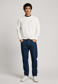 Sudadera blanca con puños acanalados, logo bordado en azul, combinada con jeans de mezclilla azul oscuro y zapatillas blancas. Ajuste casual, diseño minimalista.