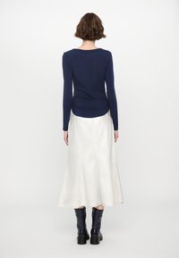 Marineblaues Langarmoberteil mit Raffung an der Taille, kombiniert mit einem weißen, fließenden Rock; Model trägt knielange marineblaue Stiefel.