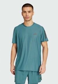 T-shirt de sport turquoise en tissu léger. Manches courtes, col rond, avec des accents à trois bandes foncées et le logo Adidas sur la poitrine.