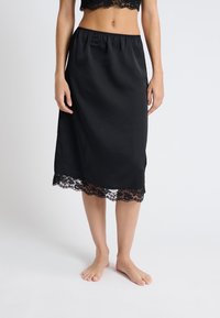 Czarna satynowa spódnica na szelkach z elastycznym pasem, z koronkowym wykończeniem u dołu i długością do kolan. Miękka faktura i gładkie wykończenie.