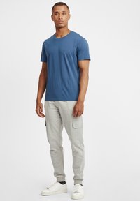 Blå crew-neck t-skjorte kombinert med lys grå cargo bukser med sidelommer og en strukturert stoff. Skoene er hvite med svarte detaljer.