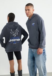 Gray hoodies with "Cosmic dreamer" prints in blue and silver swirl design, featuring front pockets and soft fabric. Styled with jeans and leggings.