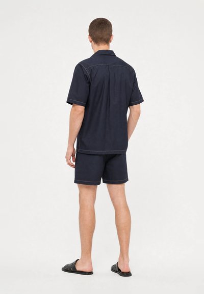 Homme debout, de dos, portant une chemise bleu marine à manches courtes et un short assorti avec des sandales noires sur un fond uni.