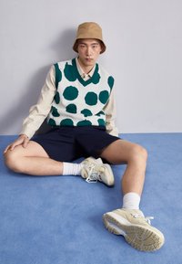 Zeleno-bílé puntíkované vestičce přes béžovou košili, námořnické šortky a krémové tenisky s modrými akcenty, sedící na světle modrém povrchu.