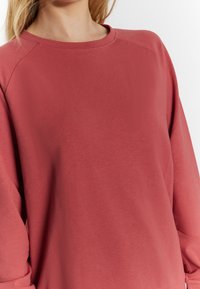 Detailní záběr na osobu, která má na sobě dlouhý rukáv, top v barvě prachové růže s kulatým výstřihem na pozadí bílé barvy.