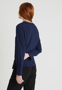 Blouse bleu marine à manches longues en tissu lisse, col rond et poignets boutonnés, associée à un pantalon noir fuselé.