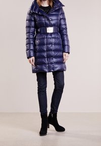 Marineblå dynejakke med bælte i taljen, designet med høj krave, quiltet tekstur og sølvfarvet hardware. Bæres over mørke, tætsiddende jeans.