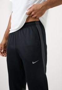 Mężczyzna podnoszący białą koszulę, aby odsłonić talię, ubrany w czarne spodnie dresowe Nike z bocznymi paskami i widocznym logo swoosh na jasnym tle.