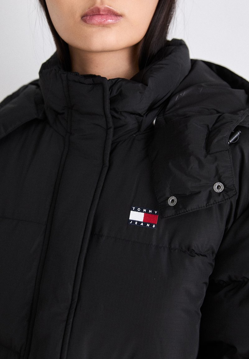 Schwarze Daunenjacke mit hohem Kragen und Druckknopfverschluss. Mit einem auffälligen, gestickten Tommy Jeans-Logo in Rot, Weiß und Blau.