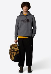 Sudadera gris con logo, camiseta amarilla por debajo, pantalones negros con bolsillos, mochila negra y amarilla con estampado, zapatos marrones con cordones.