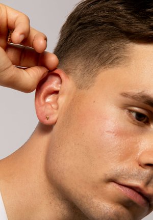 Ein goldener Blitzohrring im linken Ohr eines Mannes, gehalten von einer Hand. Kurze Haare und glatte Haut sind im Close-up sichtbar.