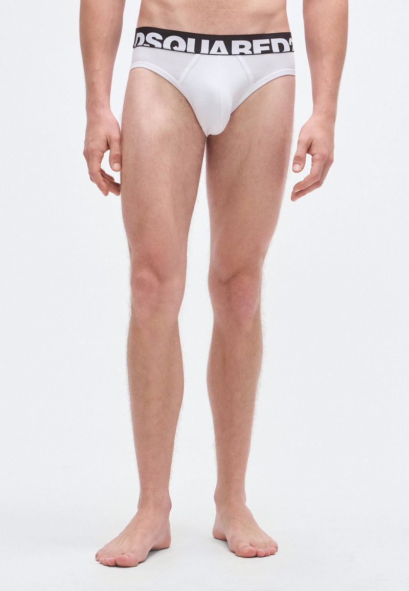Braguitas blancas con una cintura negra con el texto "DSQUARED2". La tela parece lisa y el diseño es ajustado. Las piernas están expuestas, mostrando la textura natural de la piel.
