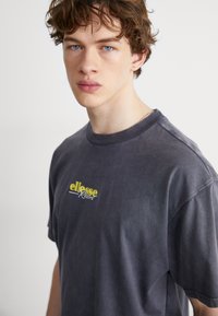 Pelēka kokvilnas T-kreklu ar īsām piedurknēm, ar dzelteno "ellesse Resort" logo uz krūtīm. Audums izskatās mīksts un nedaudz nolietots.