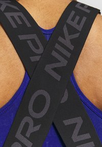 Czarne sportowe paski z szarym napisem "NIKE PRO" krzyżują się na niebieskiej koszulce bez rękawów. Projekt jest nowoczesny, z gładką fakturą i trwałymi materiałami.