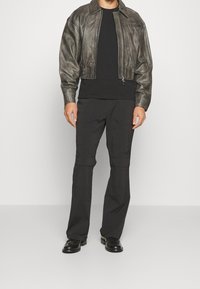 Man med kort mörkgrå läderjacka över svart skjorta, svarta byxor med knäskärningar och svarta dressade skor mot vit bakgrund.