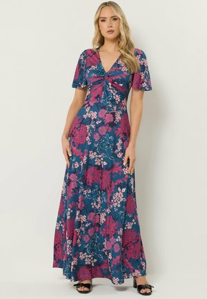 Kobieta w niebieskiej długiej sukni maxi z różowymi kwiatowymi wzorami, krótkimi rękawami, dekoltem w serek i marszczeniem na wysokości klatki piersiowej, stojąca na tle białej ściany.