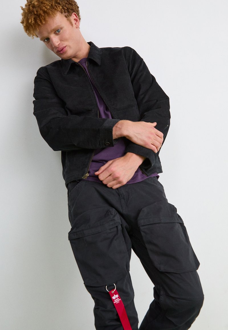 Czarna kurtka z kordurowej tkaniny z kołnierzem i zamkiem błyskawicznym, zestawiona z czarnymi spodniami cargo z wieloma kieszeniami oraz czerwoną zawieszką na klucze.