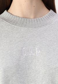 Šviesiai pilkas sportinis megztinis su išformuotu apykakle ir siuvinėtu "GAP" logotipu. Minkšta, tekstūrinė medžiaga, minimalus dizainas ir jokie matomi raštai.