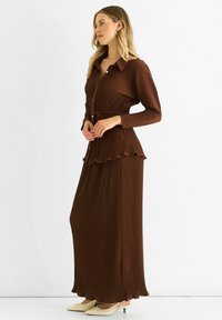 Brown pleated maxi dress with long sleeves, a collar, button front, and ruffled hem. Paired with cream slip-on shoes.