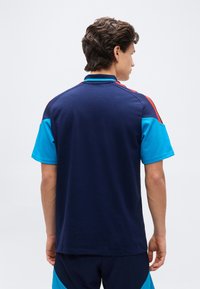 Jaunas vyras su trumpais garbanotais plaukais, dėvintis tamsiai mėlyną marškinių su turkio spalvos trumpomis rankovėmis ir raudonomis juostelėmis ant pečių, žiūrintis į šalį.