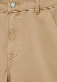 Gros plan sur un pantalon beige avec bouton visible, passant de ceinture, bord de poche et coutures détaillées sur un tissu épais.