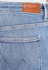 Vaqueros de mezclilla azul claro con una etiqueta de cuero de Wrangler en la parte trasera de la cintura, que presentan bolsillos estándar y un patrón de costura visible.
